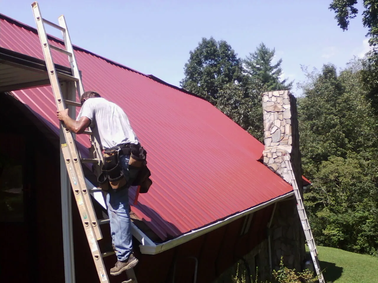 Roofer on a ladder beside a red metal roof and stone chimney — Soffit & Fascia Repair by Roofing Brothers in Duck River